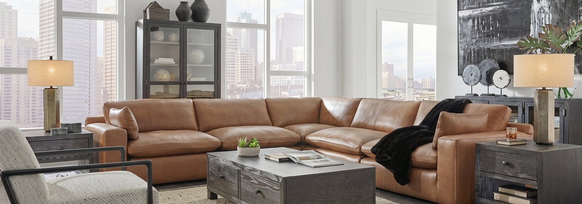 A caramel leather sectional sofa arranged in an U-shape in a modern living room with large windows, city views, side tables, lamps, and a coffee table.